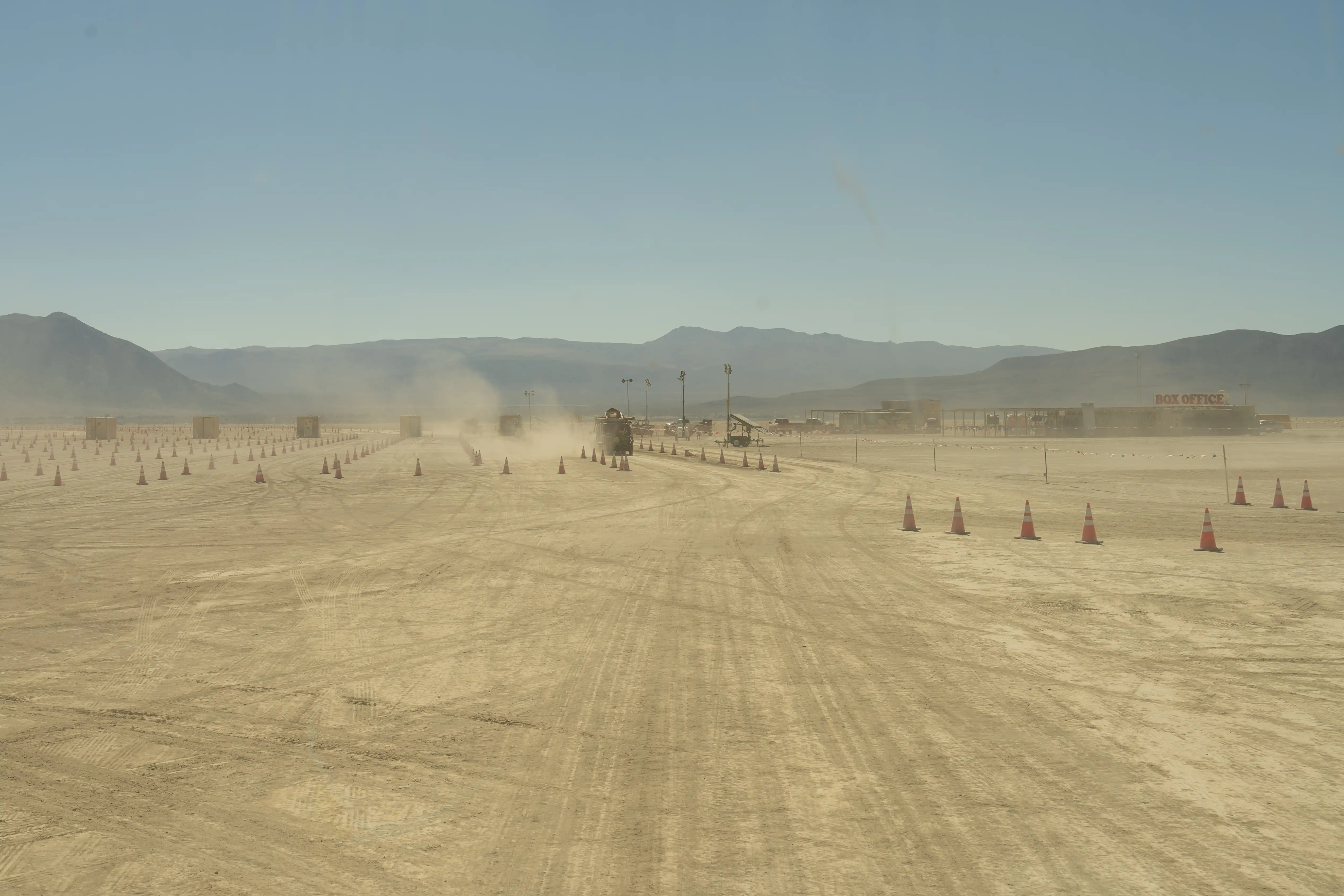 Entrance to Burning Man. A dusty, dry lake bed with mountains on the horizon. Traffic cones have been arranged to create lanes. Trucks entering are kicking up dust. A structure with a sign that says Box Office.