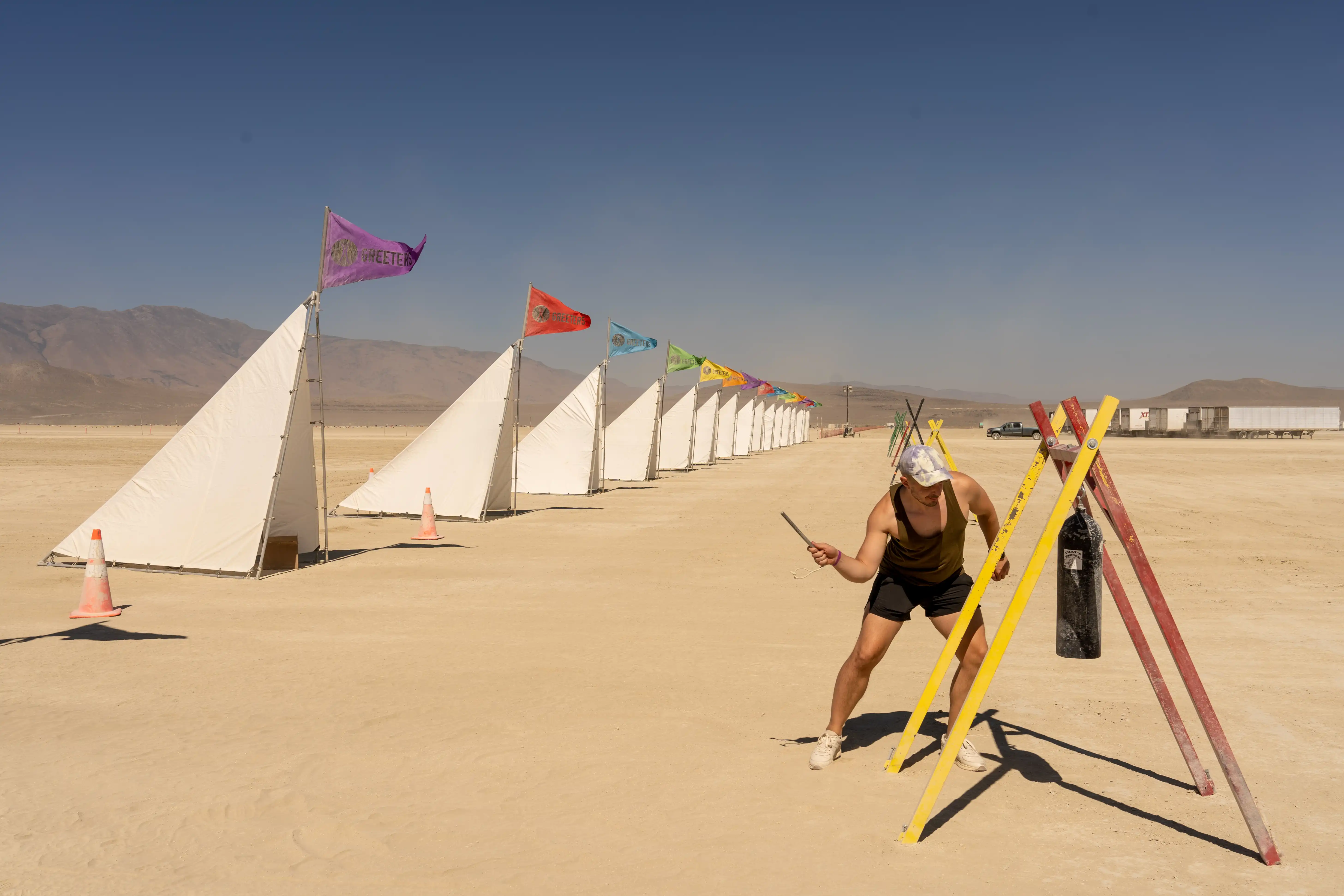 Greeters station at Burning Man. In the foreground, a muscular man in a tank-top is about to hit a large cylindrical bell. In the background, a row of white, triangular lean-to tents with colorful flags labeled Greeters.