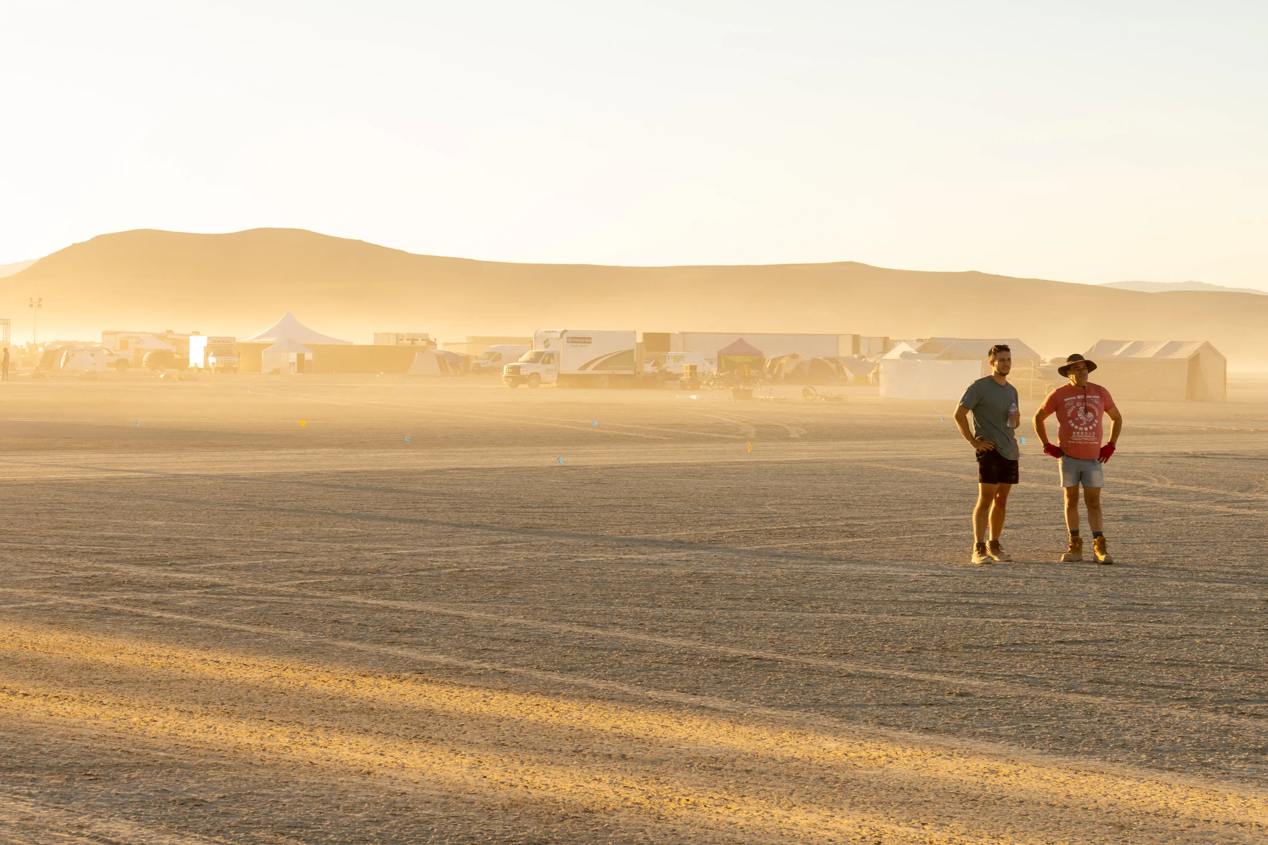 Dry lake bed of Burning Man playa in the foreground, mountains in the background. Two men with arms akimbo inspect a big, empty stretch of desert. Behind them a hazy, dusty scene of tents, trucks, and shade structures.