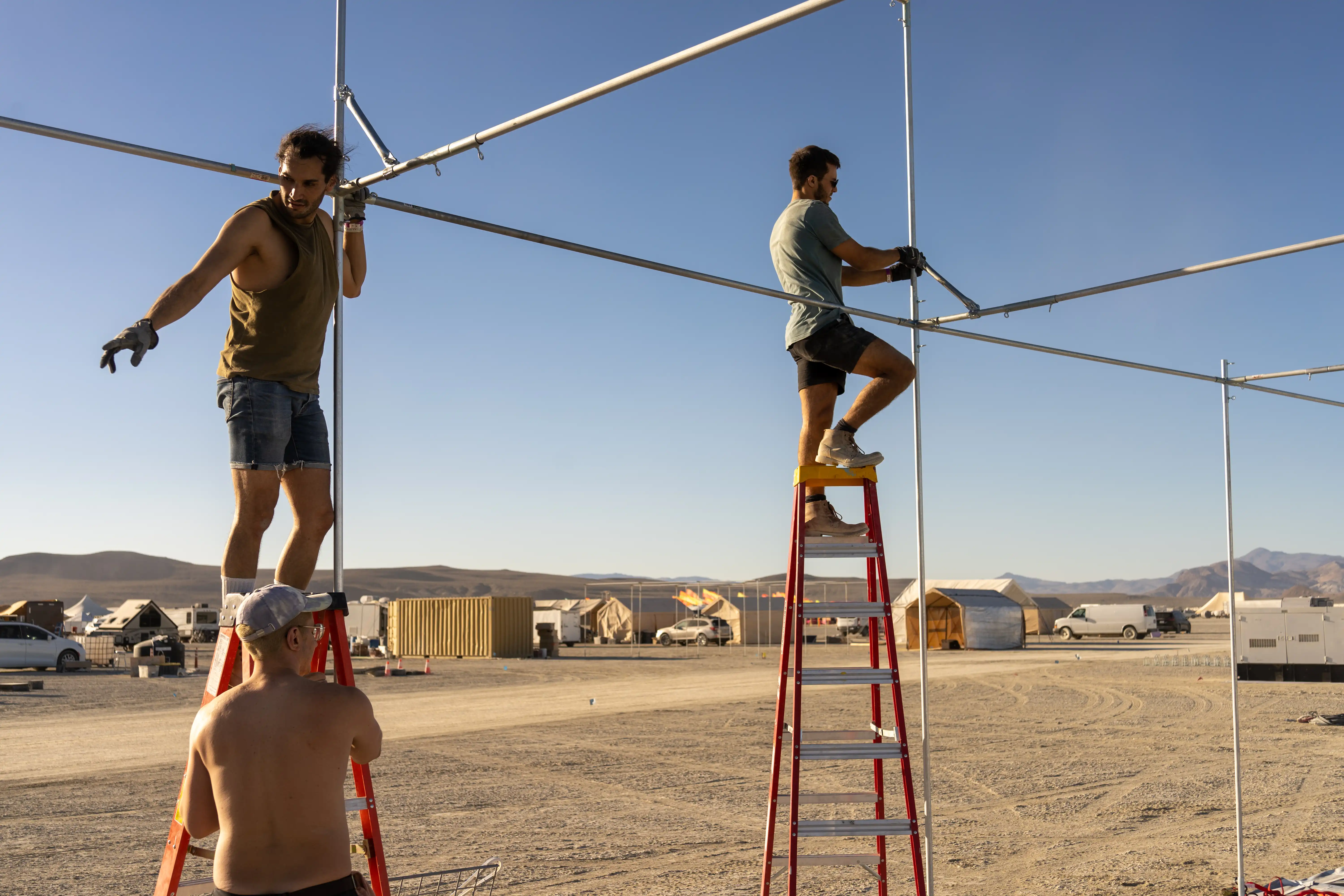 Building a shade struture at Burning Man. Two men are on ladders while another shirtless man holds the ladder. The men are erecting the frame of a structure by connecting metal pipes. In the background, other camps, tents, and vehicles.