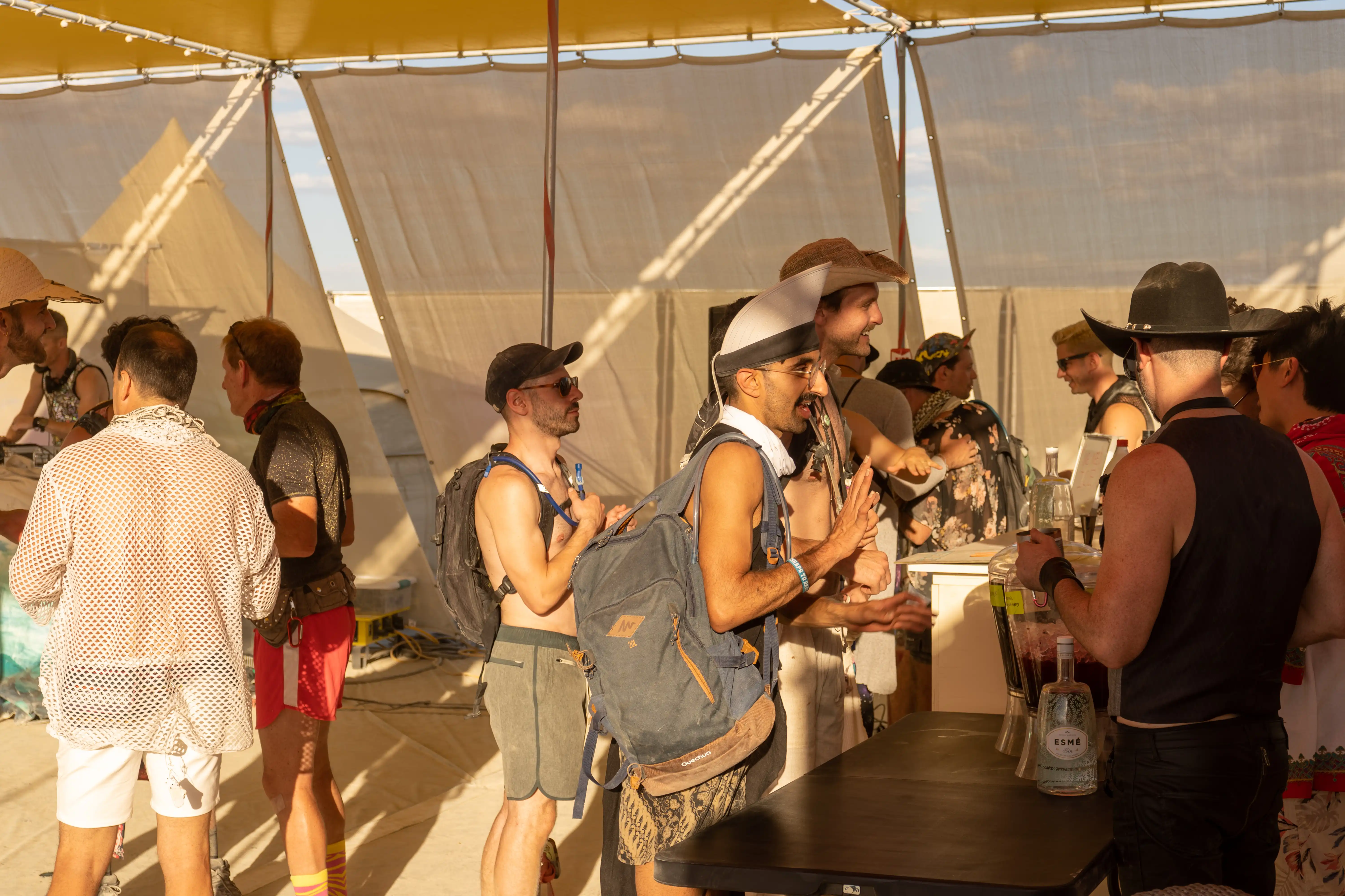 Sunset. The bar at the Future Turtles theme camp. Inside the shade structure, with tarps above and sloped walls made of white mesh to deflect wind. A DJ plays while several smiling Burners stand around at an informal, dusty bar. Bartenders are preparing drinks.
