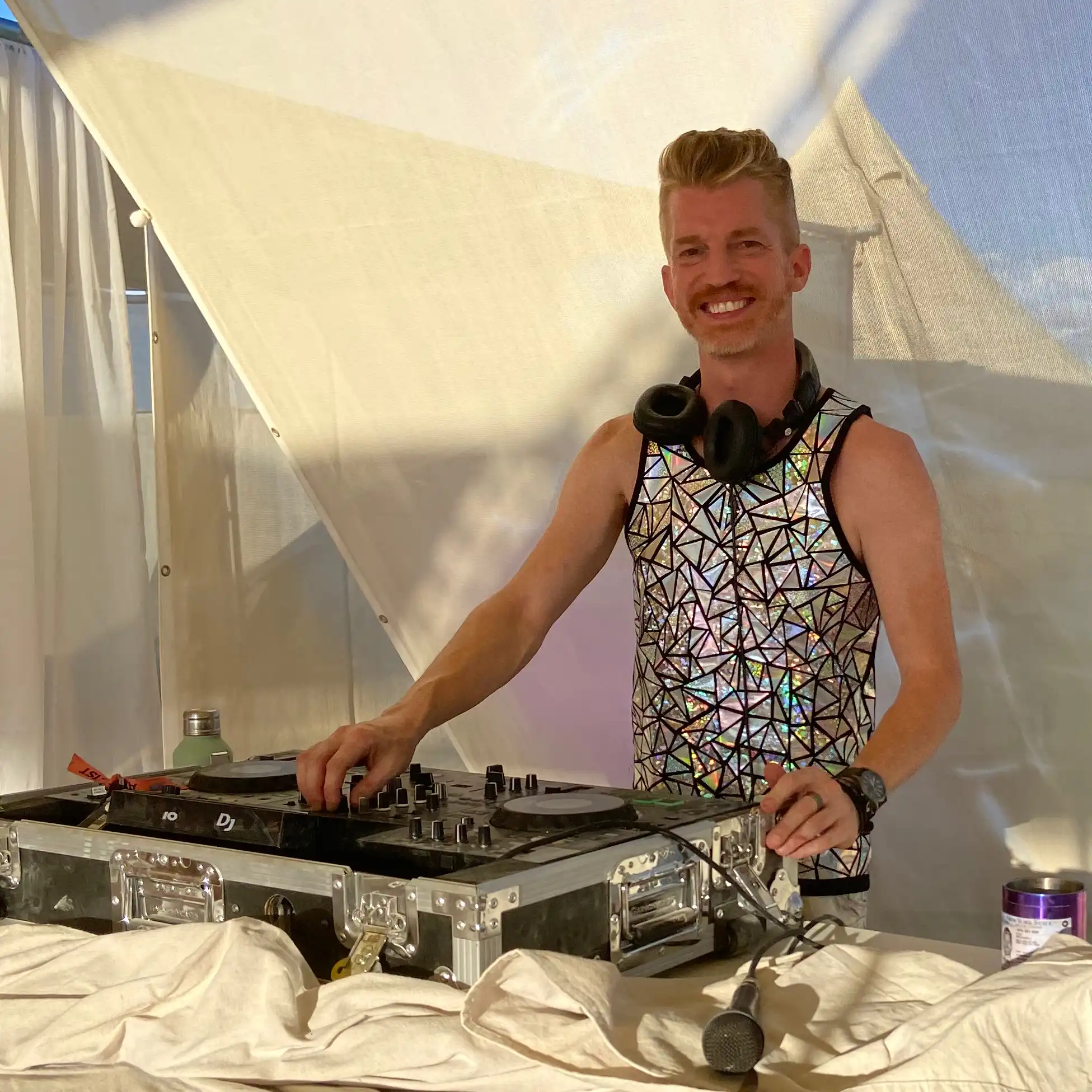 A disc jockey plays music and smiles for the camera. He is wearing a sleeveless, shiny metallic space age tank top made up of small flourescent triangles, a chunky camping watch and headphones around his neck. Behind him you can see a bell-shaped tent.