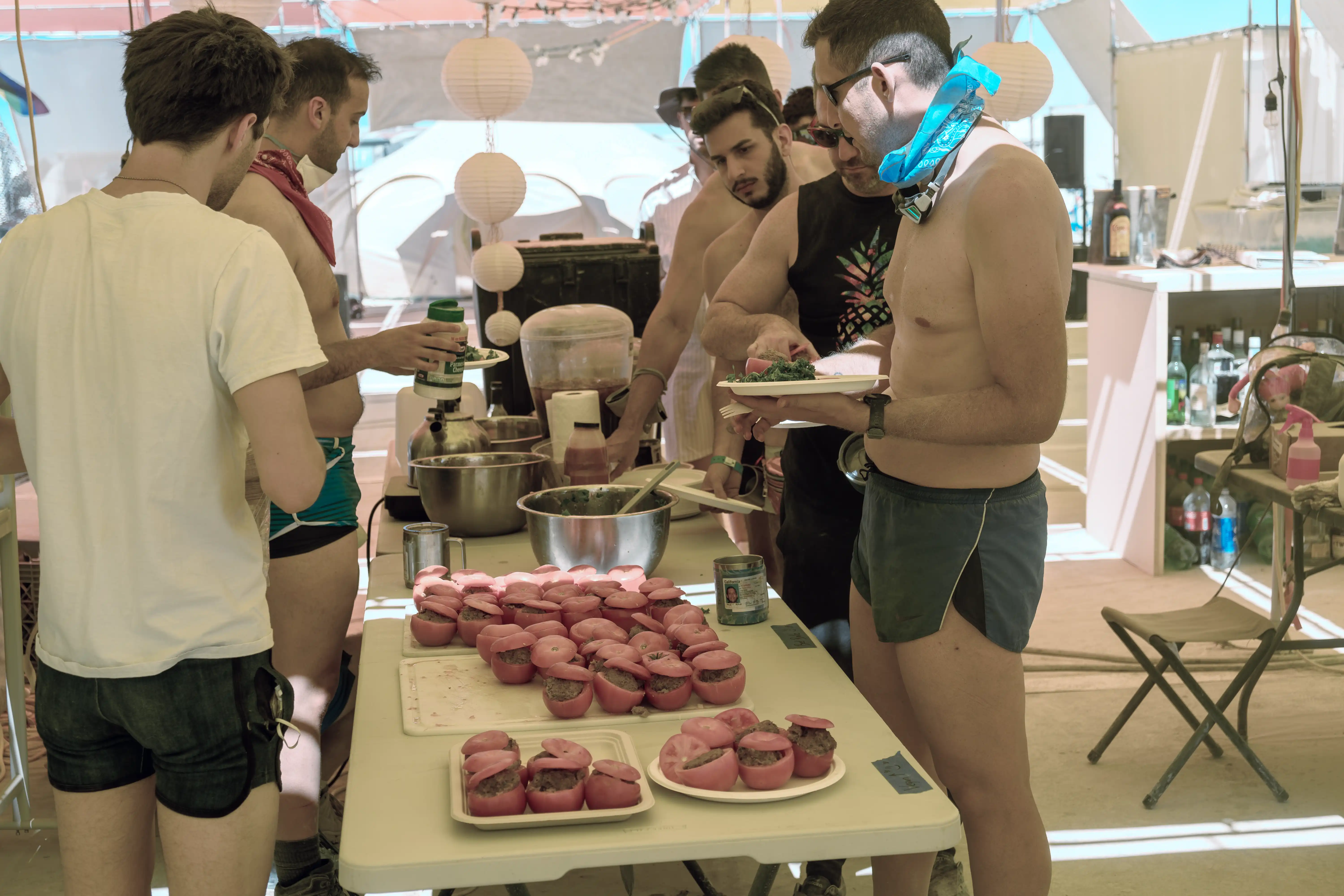 The line to get food inside the main shade at the Future Turtles. A half dozen attractive men, with and without shirts, are filling up their plates from a buffet set up on folding tables. In the foreground, tomatoes stuffed with ground beef. There are several bowls of salads and a clear urn of iced coffee.