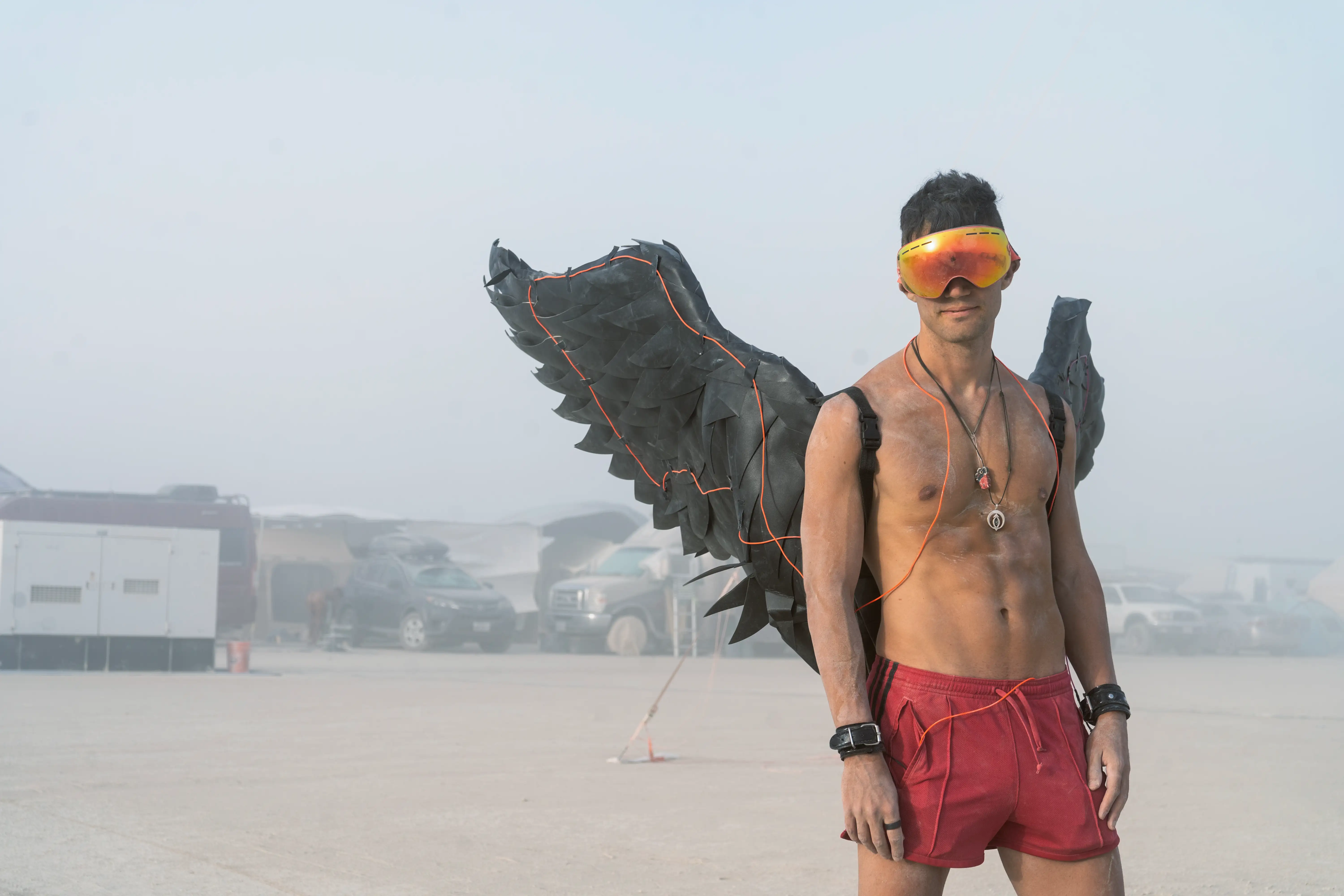 A muscular, shirtless man covered in dust in the desert of Burning Man while visibility is very low due to dust whiteouts. He is wearing giant black wings with El-wire for illumation, ski goggles, several necklaces and bracelets, and shorts.