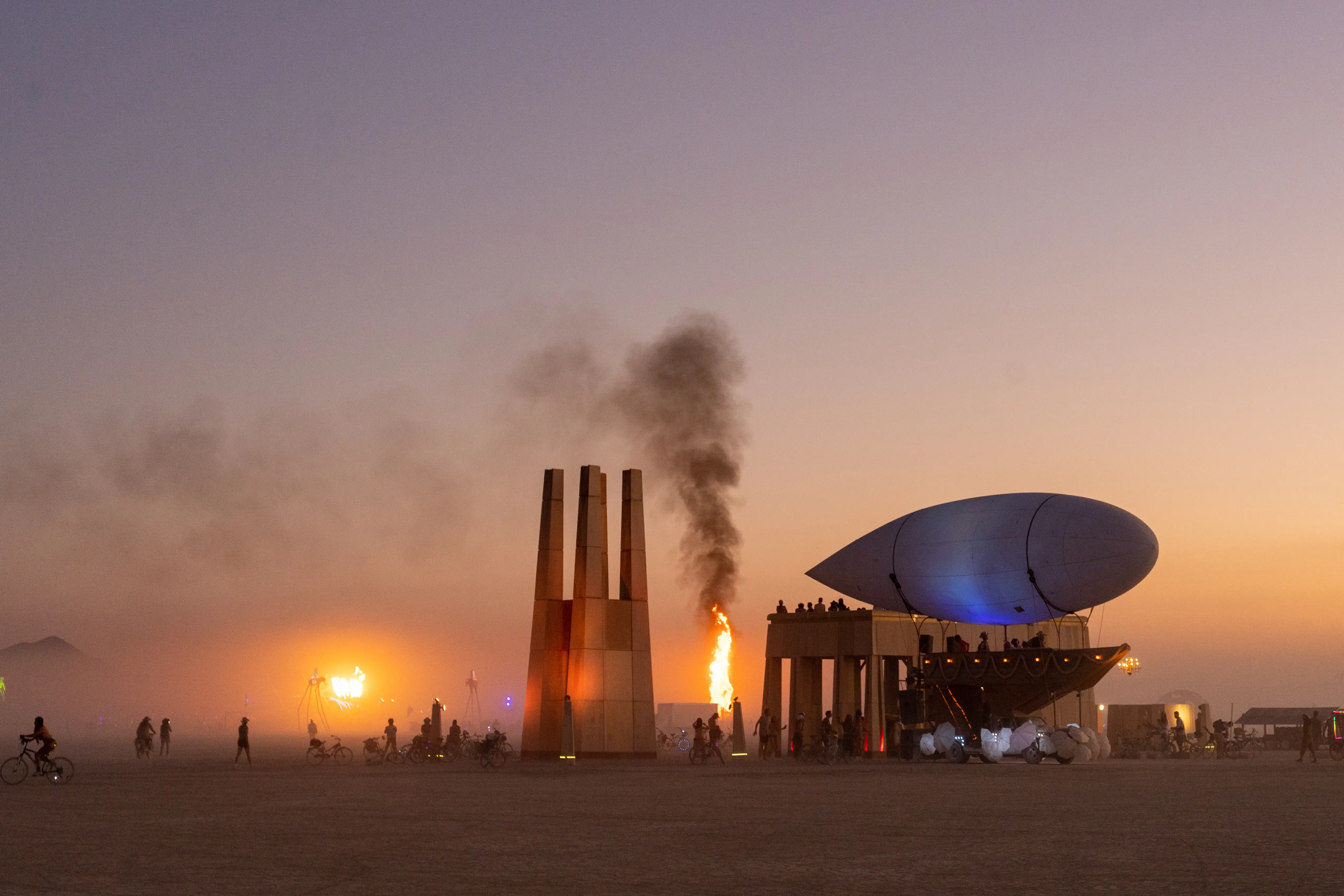 A sunset scene from the desert at Burning Man. There are two major fires in the background. In the foreground, two large buildings in the style of Afro-futurism; a tower about 40 feet high and a squat parthenon-shaped building about 25 feet high with a dozen people standing on the roof looking out at the fire. In the foreground, a mutant vehicle named Airpusher. The base of the vehicle looks like half of a sailing ship. Instead of sails, there is a large digible-shaped balloon. Bicycles and burners are seen throughout.