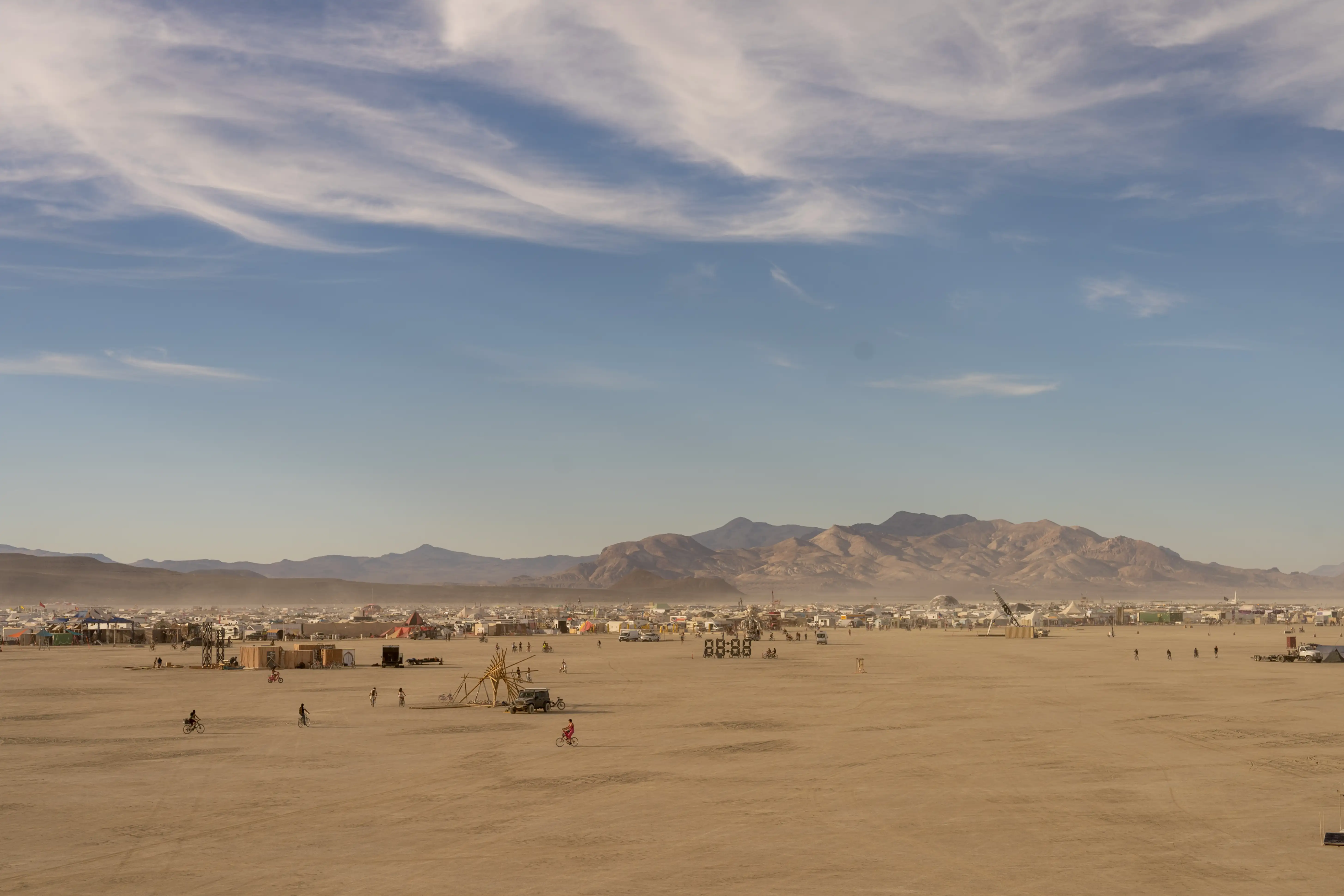 Panoramic view of the playa at Burning Man. In the foreground, the dusty desert with large sculptures sprinkled about. One structure is a digital 88:88, like a digital clock, about 10 feet tall. Burners are riding bicycles and walking around. In the midground, Black Rock City is visible in the form of tents, trucks, shade structures, and geodesic domes. In the background, the mountains. The sky is blue with a few wispy clouds.