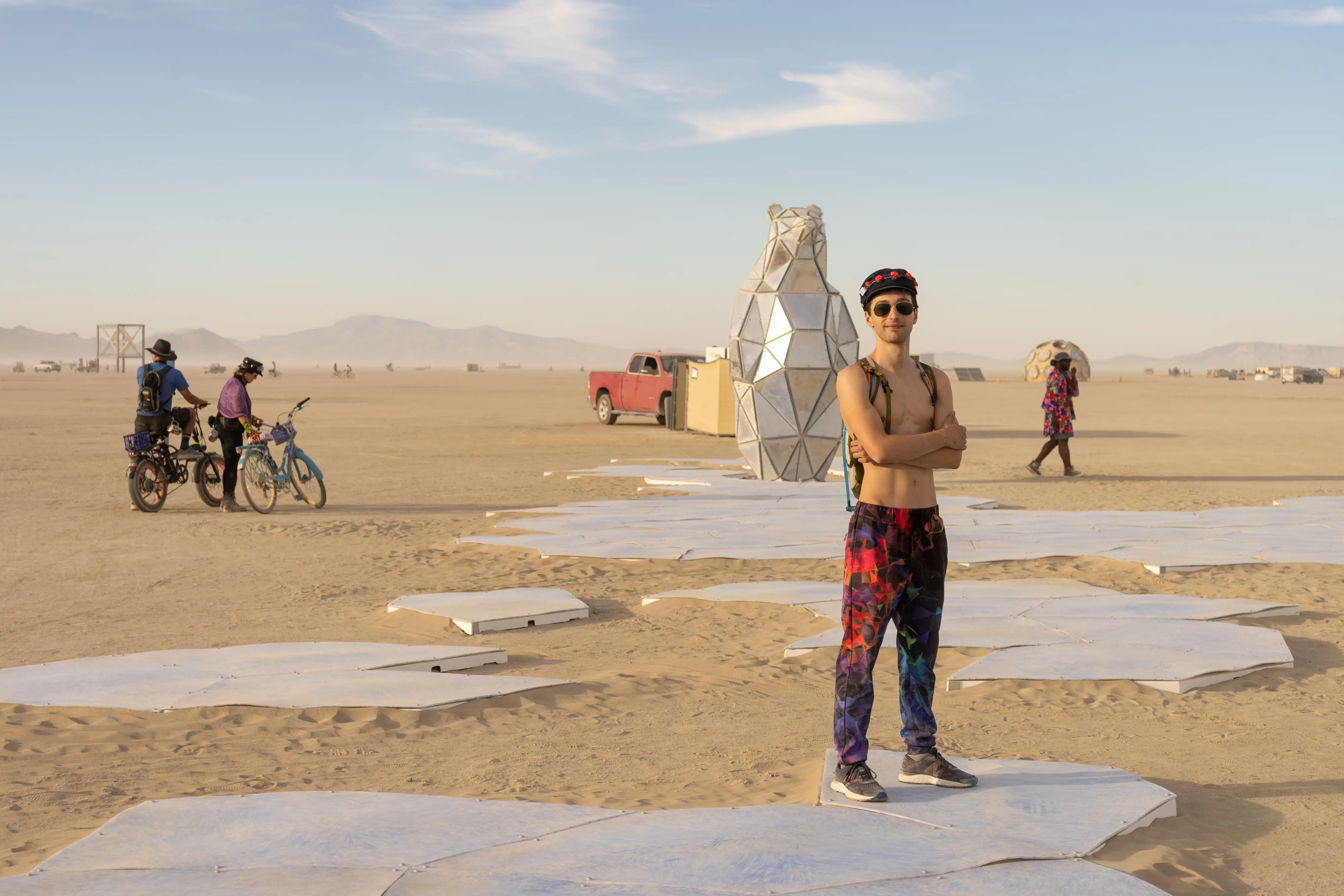 An artwork in the Black Rock Desert with a sculpture of a polar bear, made of shiny triangles, about 15 feet high. The polar bear is looking out on fragments of white shapes which represent ice floes. The ice is being swallowed by the desert, symbolizing the loss of the Polar Bear's habitat. In the foreground, a lean french twink with little-to-no body hair is wearing a beret with flowers, a backpack, sunglasses, no shirt, and colorful trousers. In the background you see other artworks, bicycles, and burners.