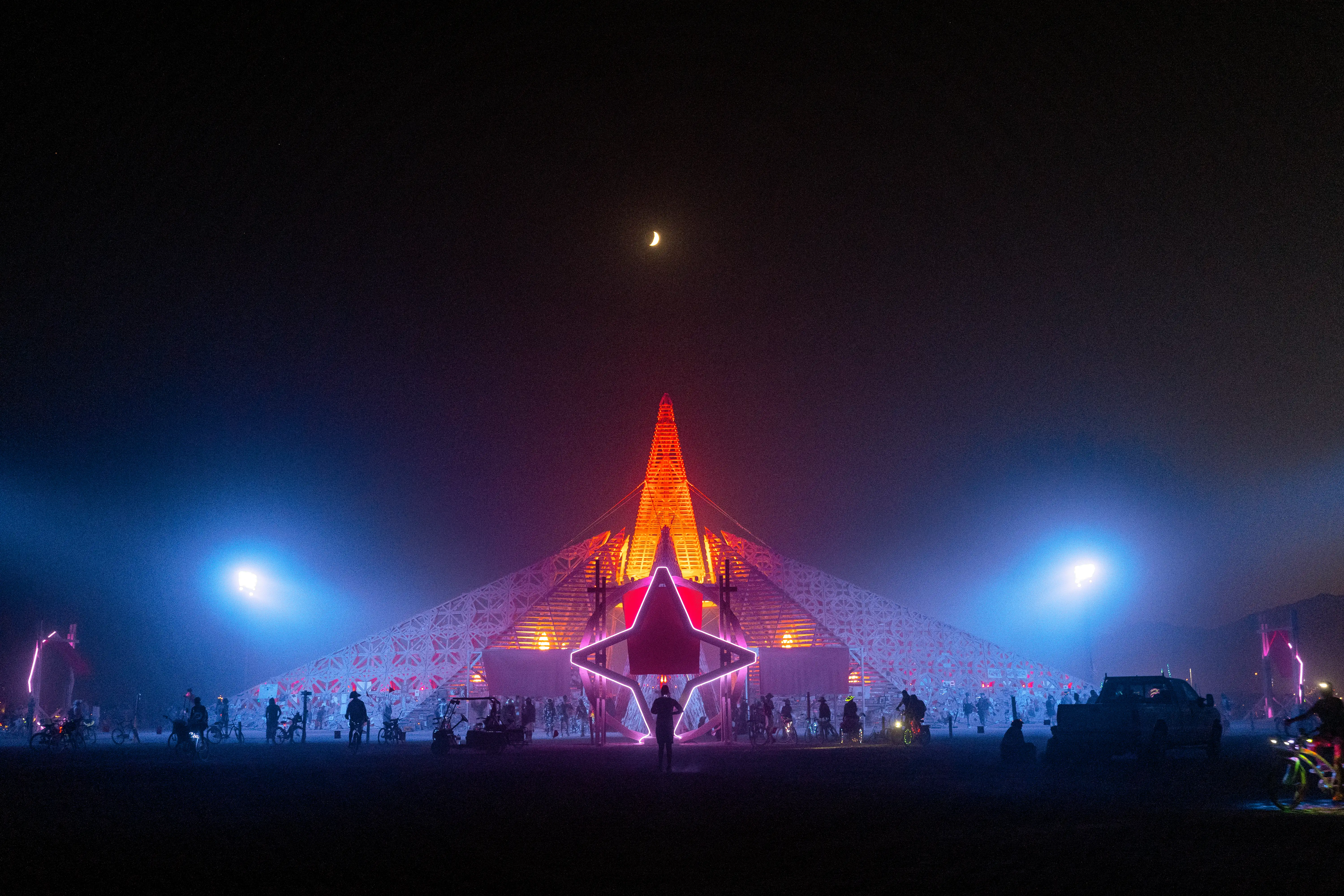 The Empyrean Temple at night, lit up dramatically. A portal in front of the temple is in the shape of a four-sided star, with a circular portal behind it. The temple is roughly pyramid shaped, with a steeper pointy tower in the center, made out of wood cut into delicate, abstract geometric patterns. The moon shines above, and burners on foot and on bicycles are seen around.