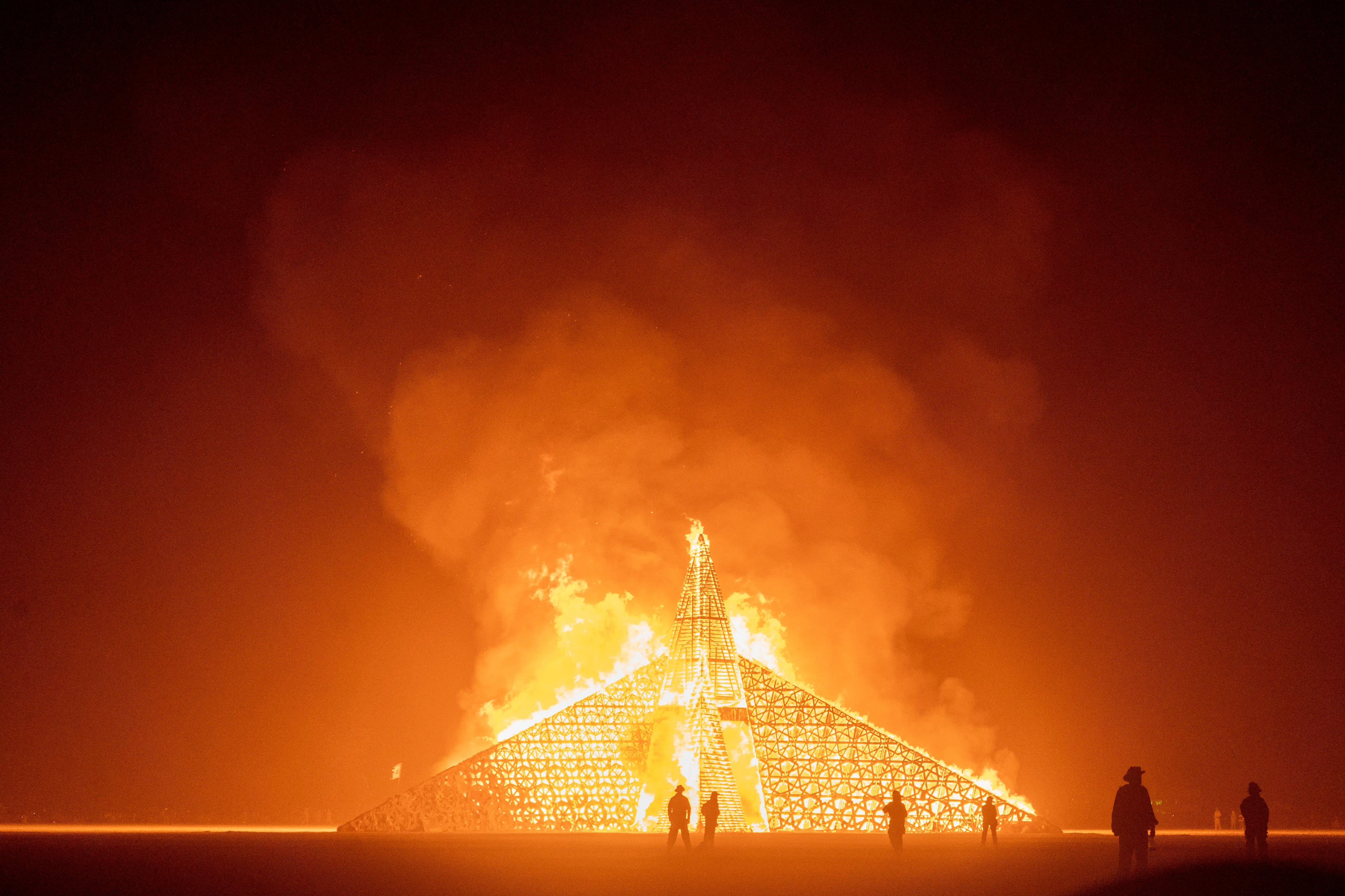 The Empyrean Temple on fire, entirely aflame. In the foreground you see silhouettes of fire safety volunteers, some in complete fireproof outfits, as the flames reach heights of 100 feet and smoke fills the sky.