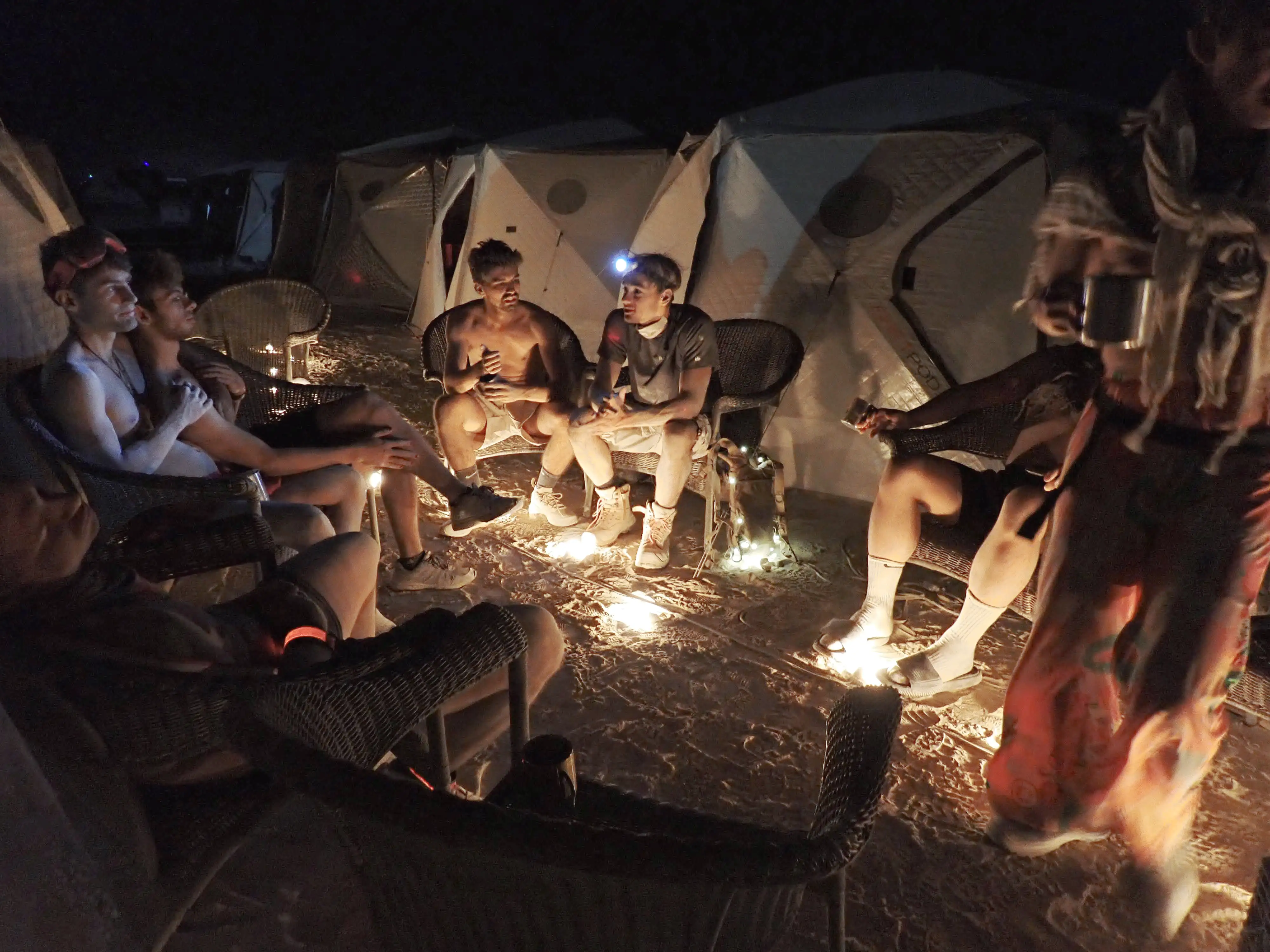 Seven people sit around on wicker chairs at night in the desert at Burning Man. One is sitting on another's lap, one is sleeping, two are chatting. There are shiftpod tents in the background, and a cozy string of lightbulbs on the desert dirt ground.