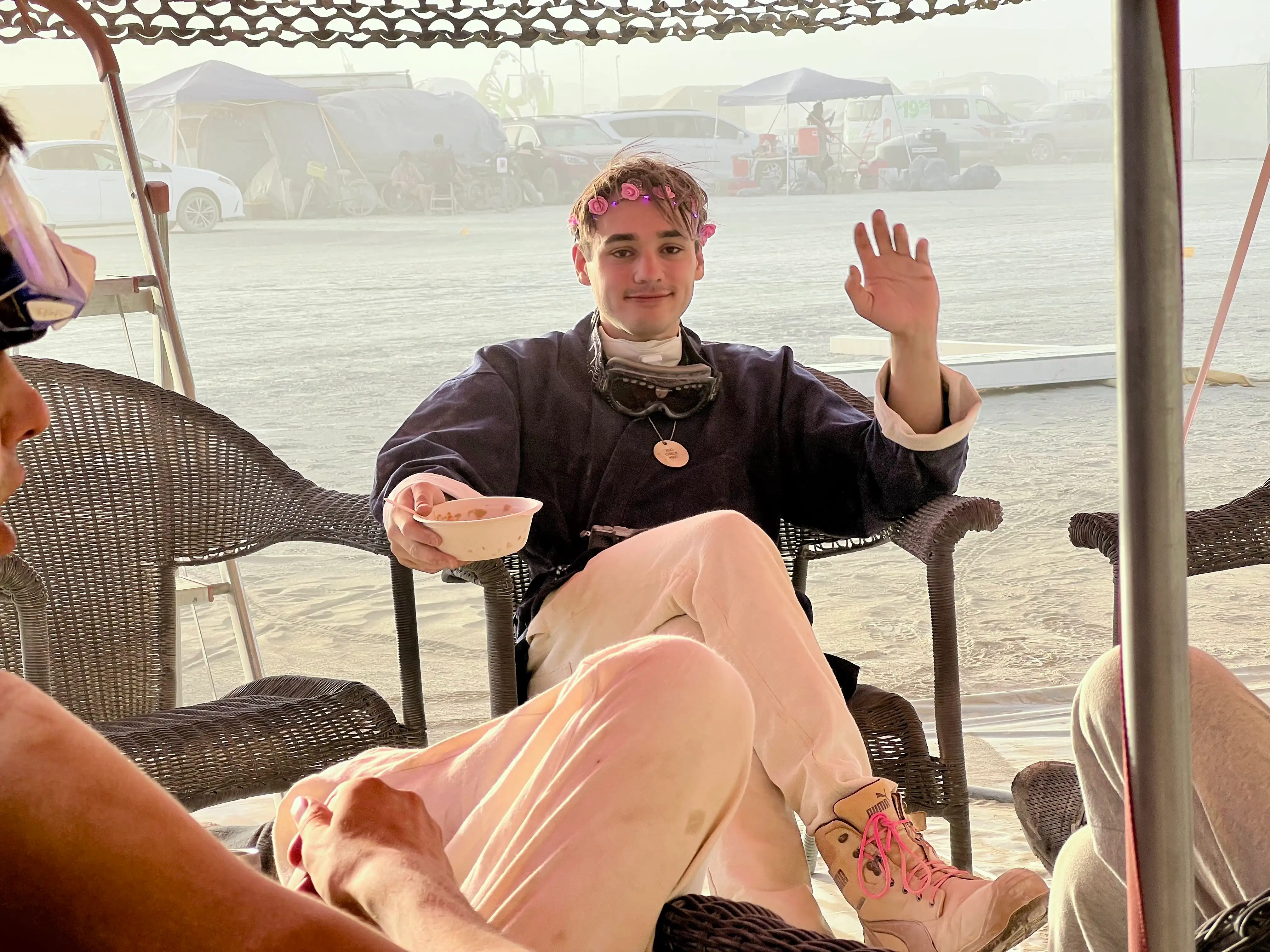 A young man sitting in the shade at the Future Turtles eating a bowl of something and waving at the camera. He is wearing an LED rose tiara, long-sleeved black shirt, white long trousers and ankle high leather boots with bright flourescent shoelaces. He has steampunk goggles and an N-95 mask around his neck and a necklace with a round pendant labeled Ego Check. In the background, it is clearly a dusty day as the camp across the street is barely visible.