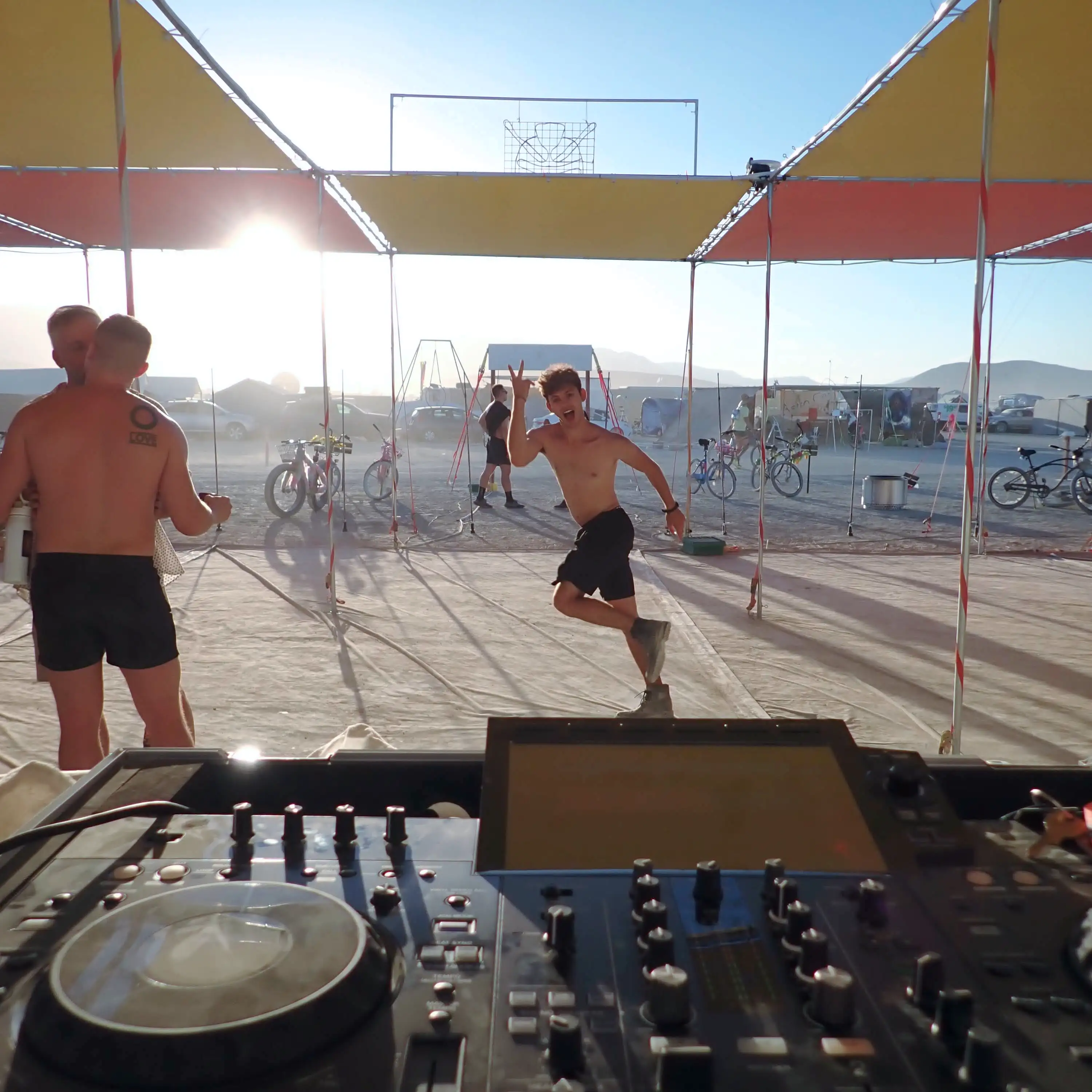 A sunset scene from the Future Turtles shade structure. Looking over the DJ equipment at a shirtless young man doing an exhuberant dance by himself. On the left side, two beefy shirtless men are making out. In the background, you can see our neighbors camp across the street where there is an aerial hoop structure set up for acrobatics.