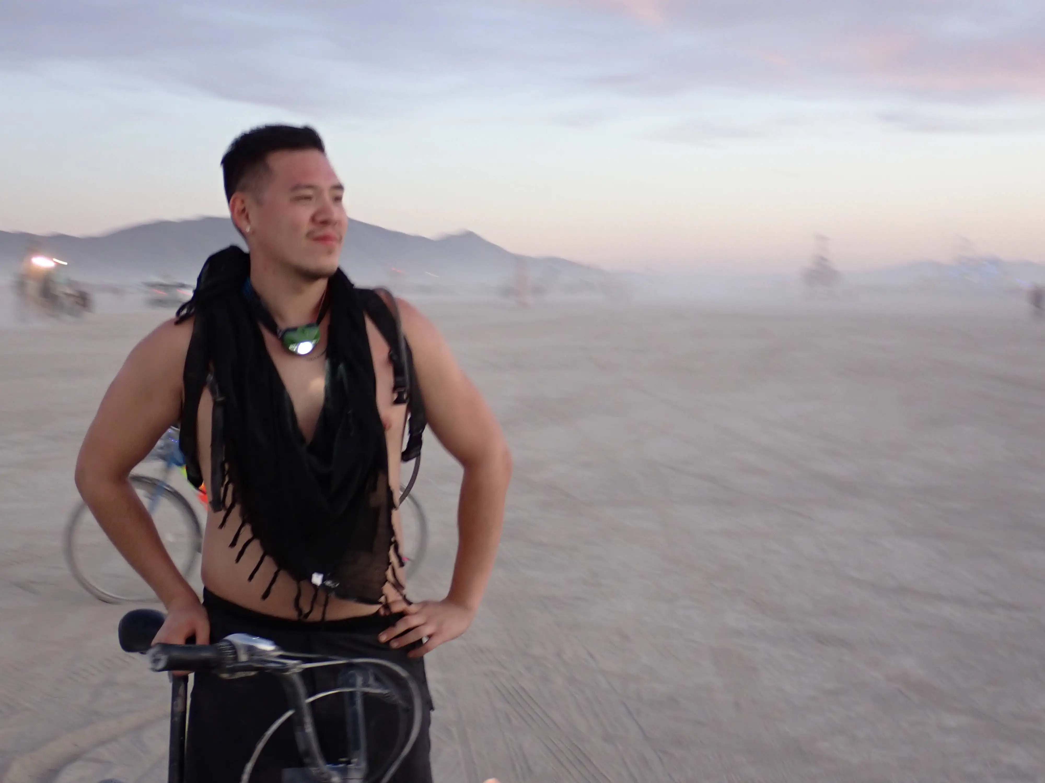 A muscular man rests on his bicycle at dawn at Burning Man. The dust makes it hard to see the background but you can make out the mountains and some mutant vehicles. He is wearing a large black scarf, no shirt, black pants, and a headlamp around his neck.