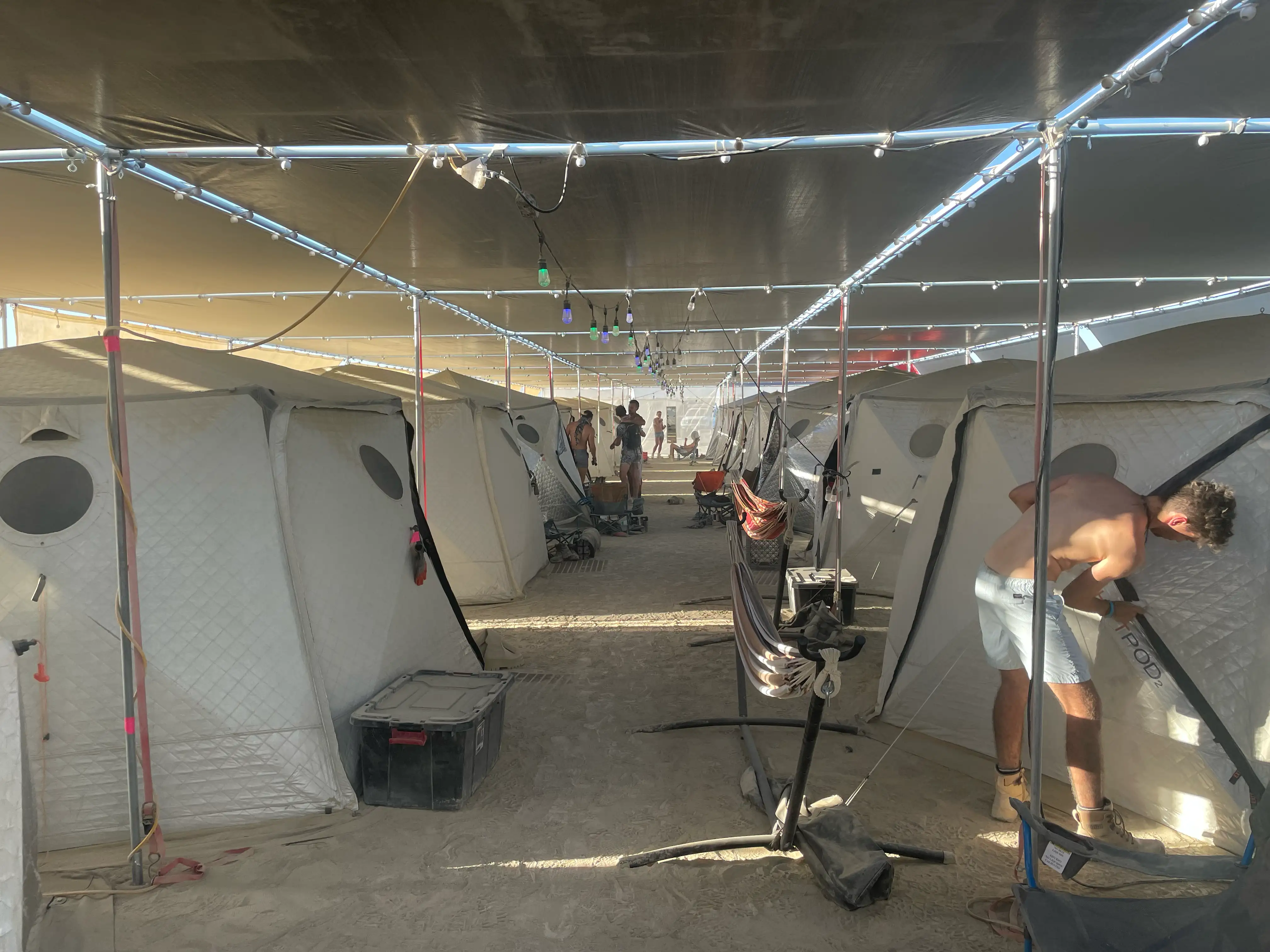 Large shade structure at Burning Man with two rows of tents and an aisle in the middle. The roof is silver tarp with a string of colored lights. Each tent is a ShiftPod 2, hexagonal shaped, just high enough to stand in and with room for two people. In the foreground, a shirtless man is unzipping the door to a tent. There are several hammocks and other camping furniture strewn around. The ground is dirt. The sun is extremely strong but the shade keeps most of it out.